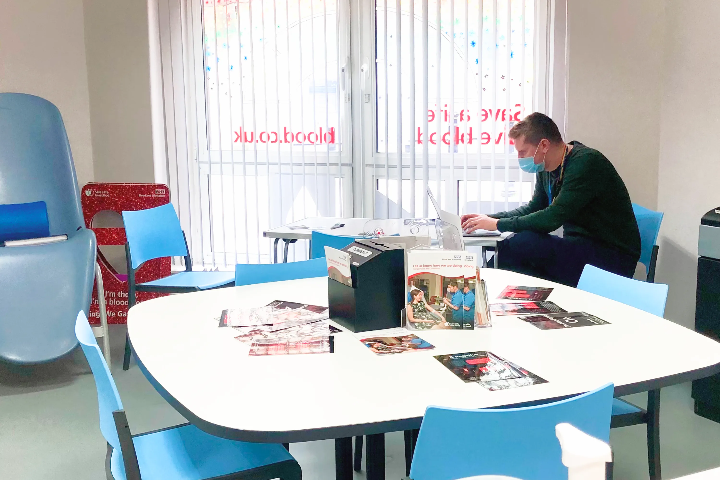 Man wearing a mask working on a laptop in an NHS blood donation waiting area with blue chairs and leaflets on a table.