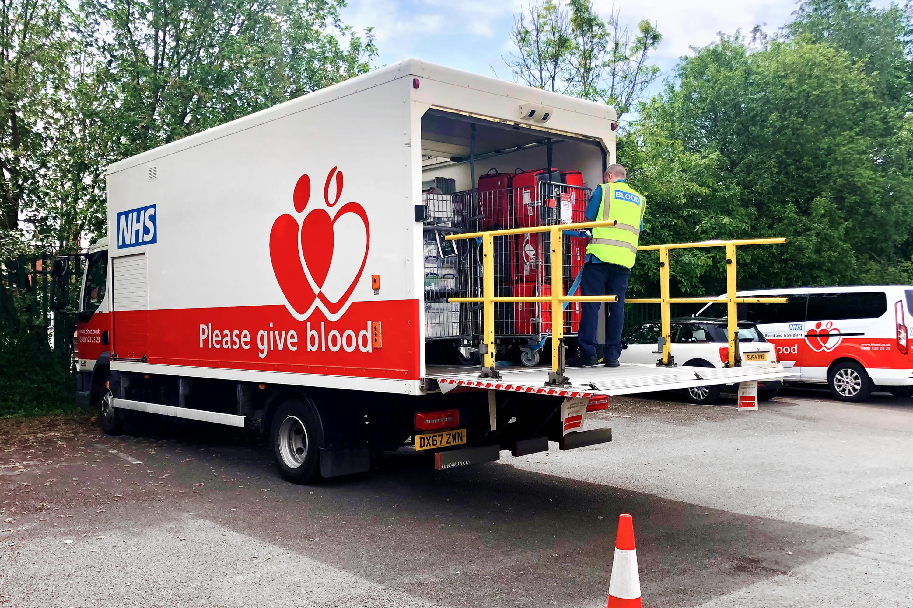 NHS Blood and Transplant lorry with “Please give blood” branding, a worker loading equipment at the back, and a support van in the background.